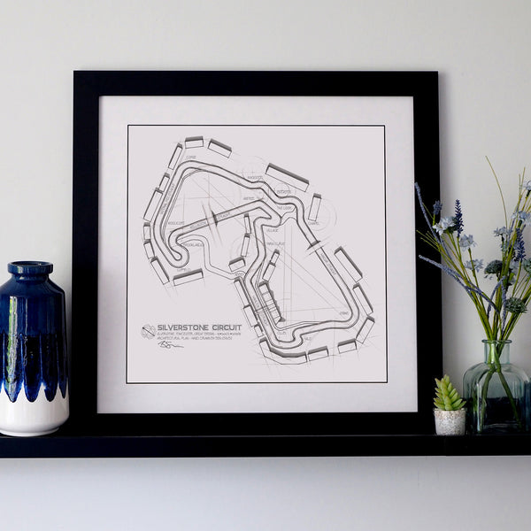 A framed black-and-white Silverstone Circuit track map print displayed on a shelf against a light gray wall, with decorative vases on both sides.
